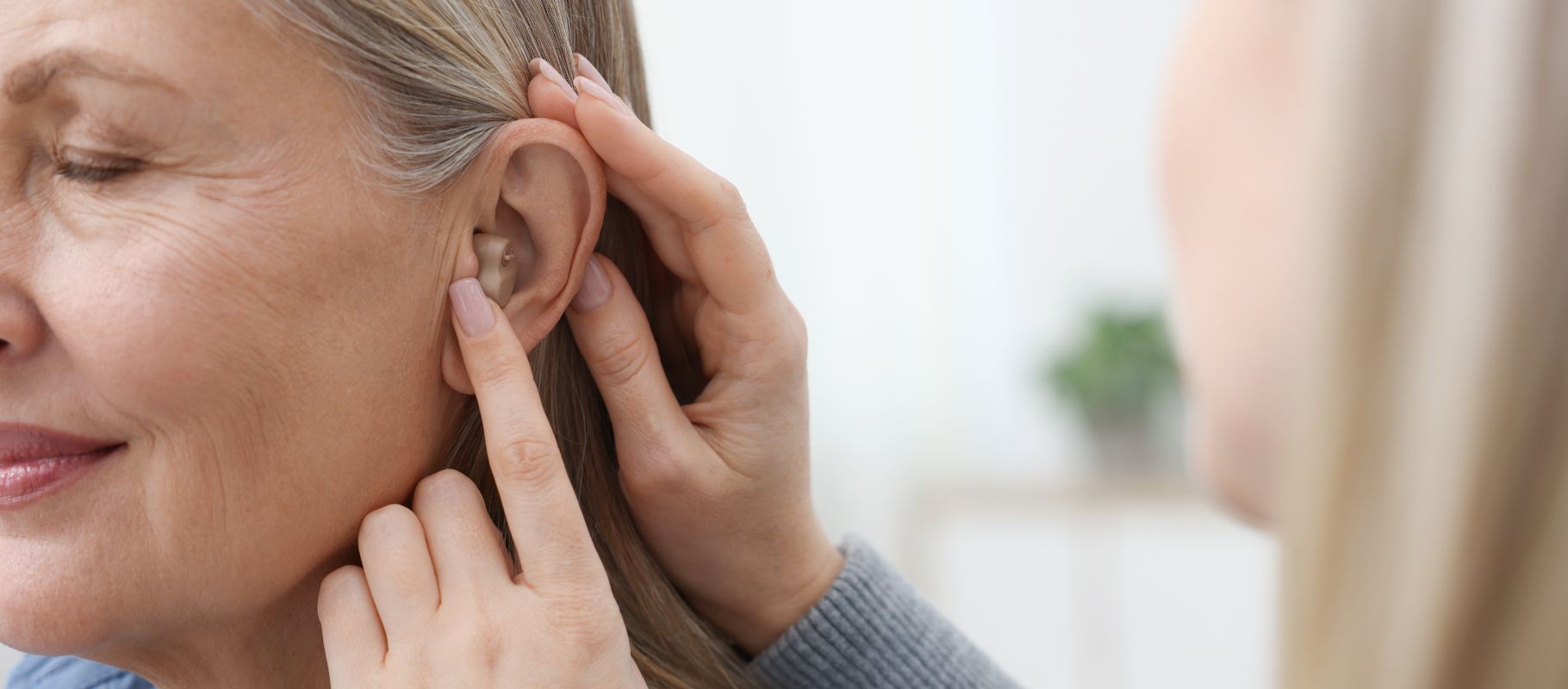 Mulher madura sorridente experimentando aparelho auditivo discreto retroauricular prata em consultório clínico moderno, paleta azul-marinho e dourado, iluminação suave, fotografia realista de produto e uso